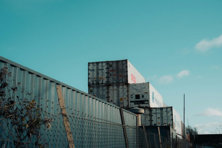 A view of stacked shipping containers beside a fence under a clear blue sky, captured outdoors.